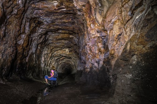 A person sits on a rock in a dimly lit cave, surrounded by textured stone walls and water on the ground.