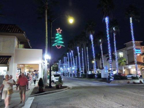 A festive street scene at night, featuring palm trees adorned with Christmas lights and a glowing tree decoration above.