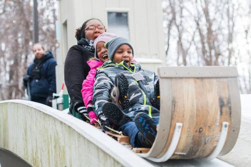A woman and two children joyfully ride a sled down a snowy hill, with trees in the background.