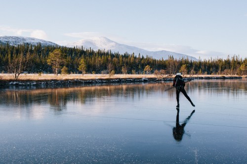 A person ice skating on a frozen lake, surrounded by snowy mountains and evergreen trees under a clear blue sky.