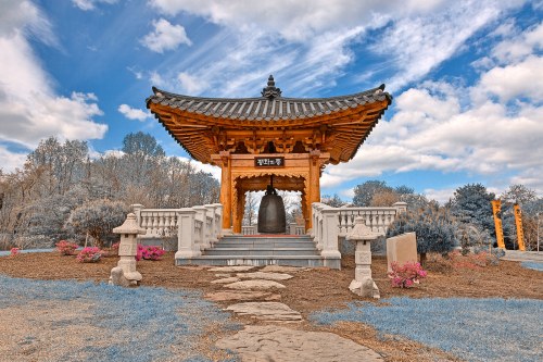 A traditional pagoda with a large bell, surrounded by colorful flowers and trees under a blue sky with clouds.