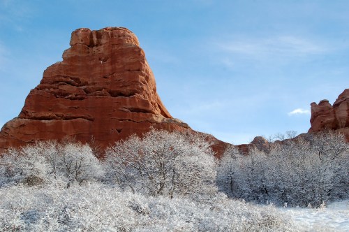 A towering red rock formation rises above a snowy landscape with frosted trees under a clear blue sky.