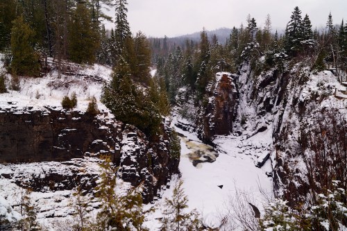 A snowy landscape featuring a river flowing through a rocky canyon, surrounded by evergreen trees and distant mountains.