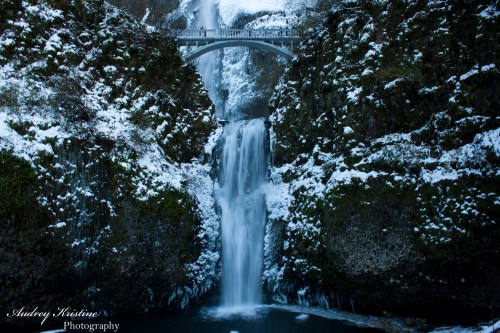 A snowy waterfall cascades down rocky cliffs, with a bridge visible above, surrounded by lush greenery.