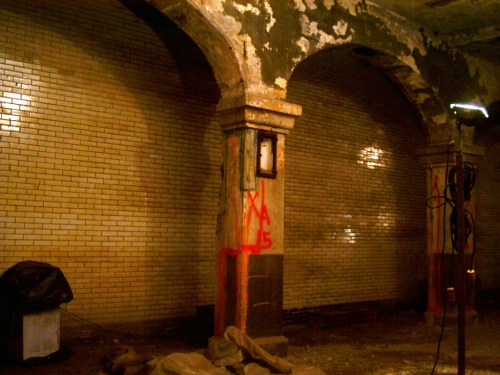 Abandoned interior with arched columns, peeling paint, and graffiti on tiled walls, dimly lit with a work light.
