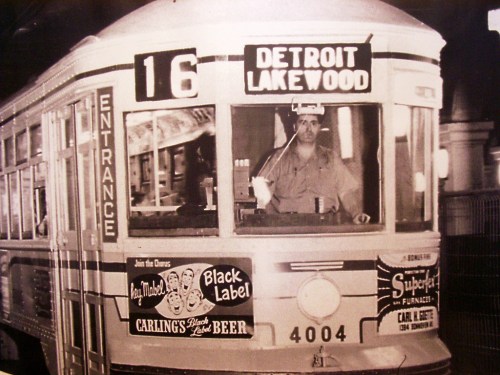 A vintage streetcar with a driver, displaying "Detroit Lakewood" and advertisements for beer and furniture.