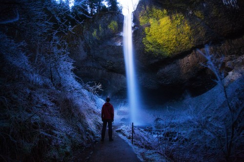 A person stands in front of a tall waterfall cascading from a rocky cliff, surrounded by frosty vegetation.