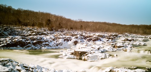 A snowy landscape featuring a flowing river and frozen waterfalls, surrounded by bare trees and rocky terrain.