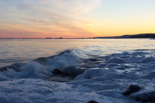 Waves crash on a rocky shore, with frothy white foam and a colorful sunset reflecting on the calm water.