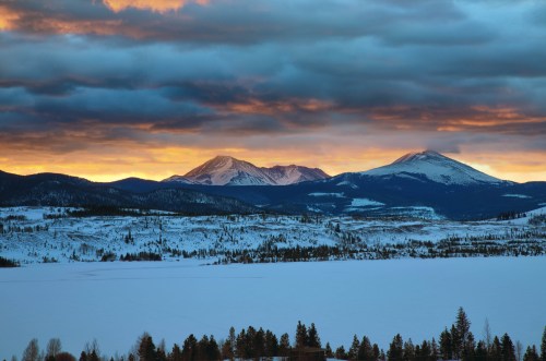 Snow-covered mountains under a dramatic sky at sunset, with a frozen lake in the foreground.