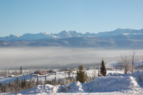 Snow-covered landscape with mountains in the background and a valley shrouded in mist under a clear blue sky.