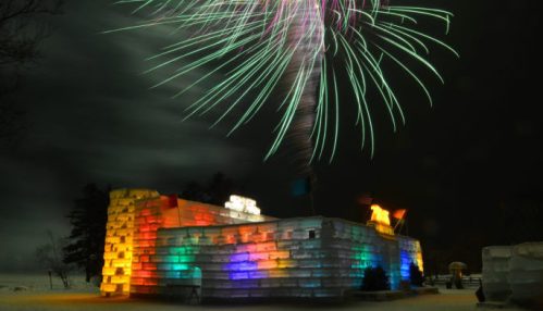 A colorful ice castle illuminated at night, with fireworks bursting above in a dark sky.