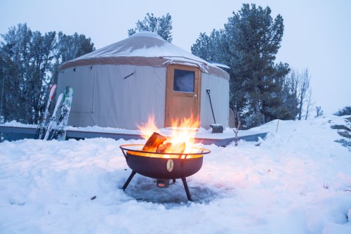 A cozy yurt in a snowy landscape, with a warm fire burning in front and ski equipment nearby.
