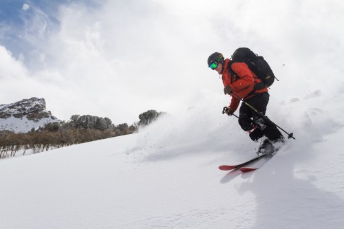 A skier in an orange jacket glides down a snowy slope, surrounded by mountains and a cloudy sky.