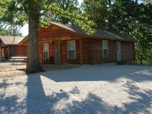 A rustic log cabin surrounded by trees, with a gravel path and porch, set in a natural outdoor setting.
