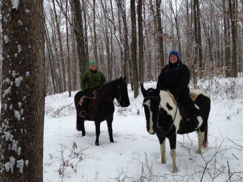 Two people on horseback in a snowy forest, surrounded by trees, wearing winter clothing.