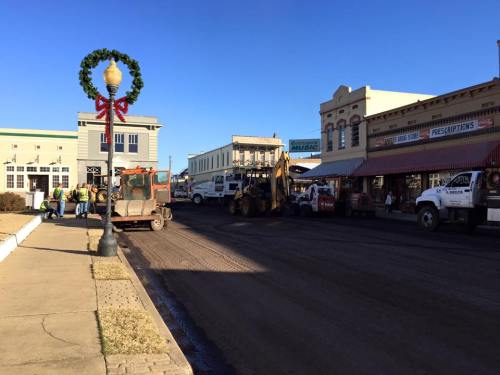 Construction vehicles and workers on a street in a small town, decorated for the holidays with a wreath.