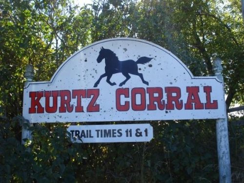 Sign for "Kurtz Corral" featuring a silhouette of a horse, with trail times listed as 11 and 1. Surrounded by greenery.