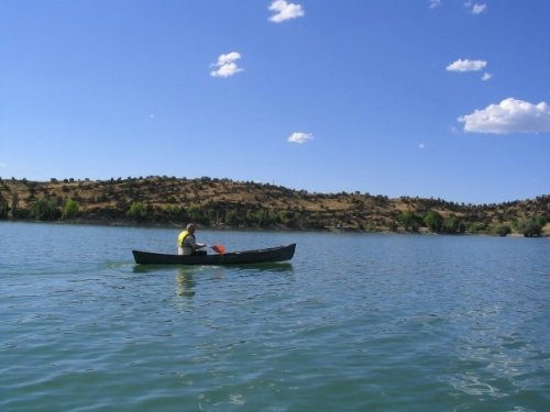A person paddles a canoe on a calm lake, surrounded by hills and a clear blue sky with scattered clouds.