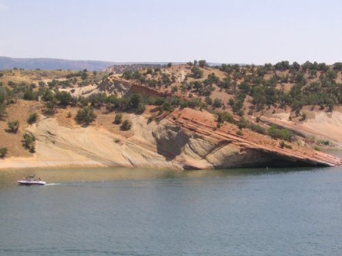 A boat glides across a calm lake, with colorful rocky cliffs and greenery lining the shore in the background.