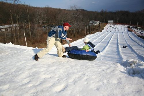 A person helps a child on a snow tubing hill, with multiple lanes of snow and trees in the background.