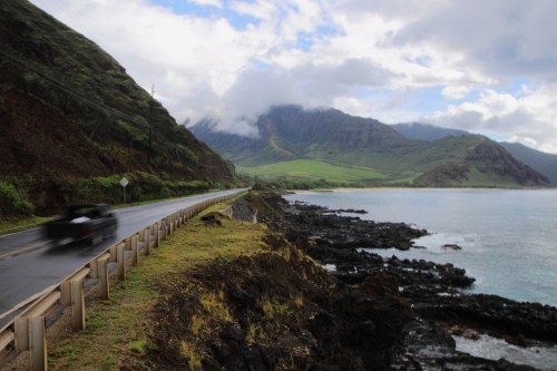 A coastal road winds along rocky shores with mountains and clouds in the background, under a partly cloudy sky.