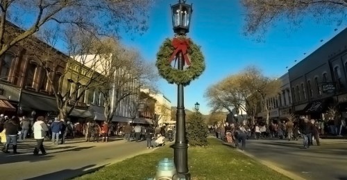 A festive street scene with a holiday wreath on a lamp post, bustling with people and lined with shops.
