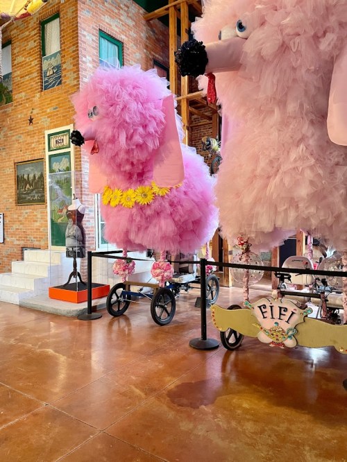 Two large, fluffy pink poodles on wheels, adorned with flowers, displayed in a colorful indoor setting.
