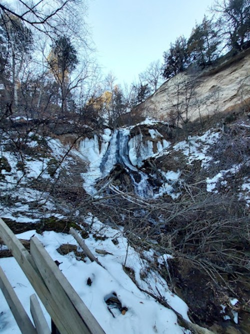 A frozen waterfall cascading down a rocky slope, surrounded by snow and bare trees.