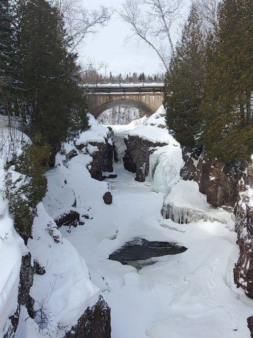 A snowy landscape featuring a frozen waterfall and a bridge above, surrounded by trees and rocky cliffs.
