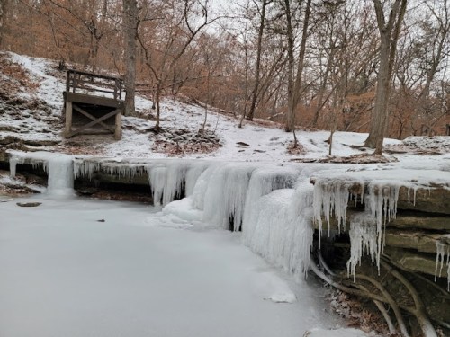 A frozen waterfall with icicles, surrounded by trees and a snowy path in a winter landscape.