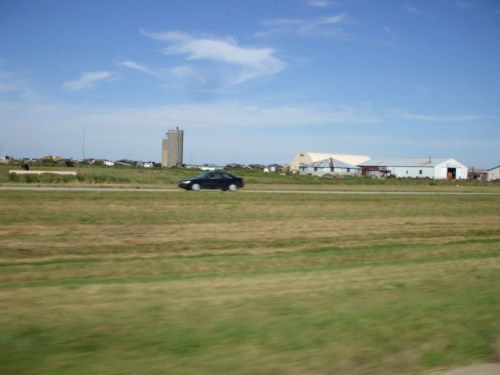 A black car drives past a rural landscape with fields, a grain silo, and several farm buildings under a blue sky.