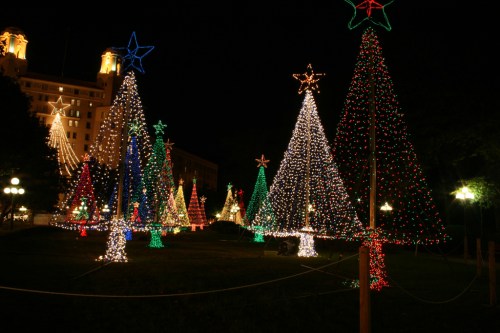 Colorful Christmas trees adorned with lights in a festive park at night, with a building illuminated in the background.