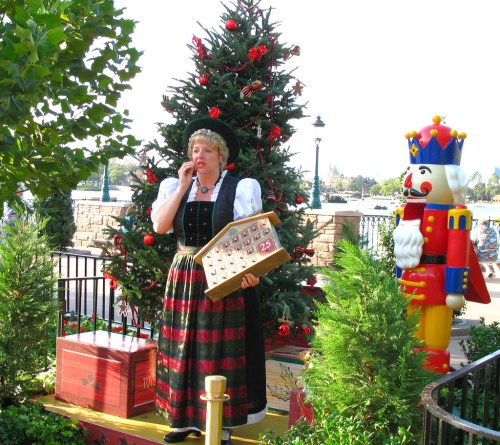 A woman in traditional attire stands by a Christmas tree, holding an advent calendar, with a nutcracker and king statue nearby.