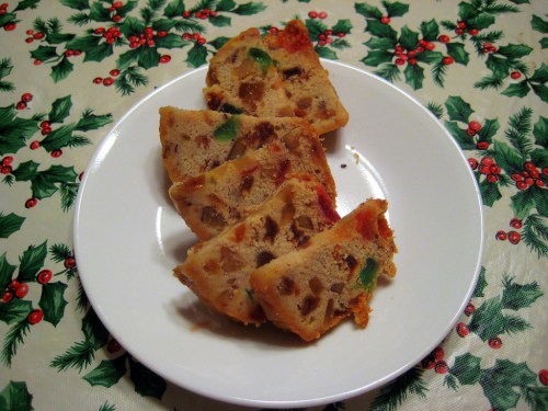 A plate of sliced fruitcake on a festive holiday tablecloth with holly and berries.