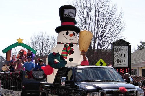 A festive parade scene featuring a large snowman float, with people celebrating and a bank sign in the background.