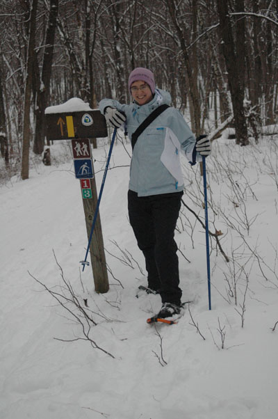 A person in winter gear stands next to a trail sign in a snowy forest, holding snowshoes and trekking poles.