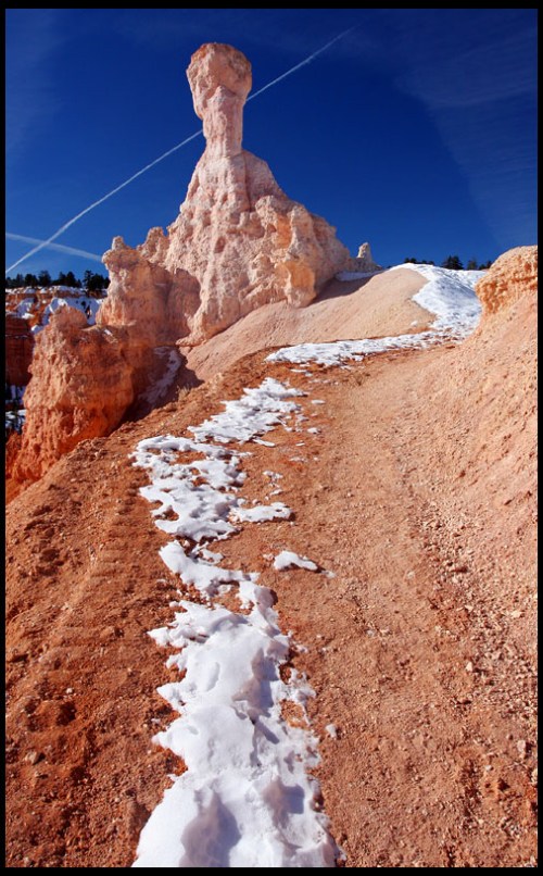 A snow-covered path winds through colorful rock formations under a clear blue sky.