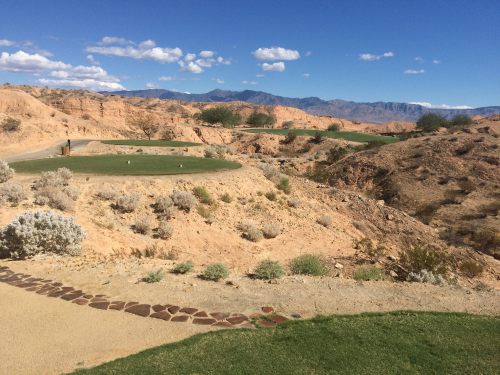 A scenic view of a golf course surrounded by rocky terrain and mountains under a clear blue sky.