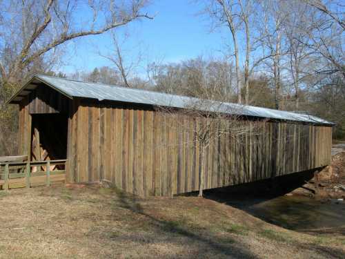 A wooden covered bridge with a metal roof, surrounded by trees and grass, spanning a small stream.