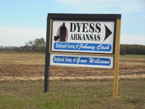 Sign for Dyess, Arkansas, indicating the boyhood homes of Johnny Cash and Gene Williams, with a rural landscape in the background.