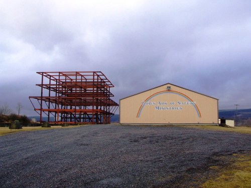 A large metal frame structure next to a building with a sign reading "God's Ark of Safety Ministries" under a cloudy sky.