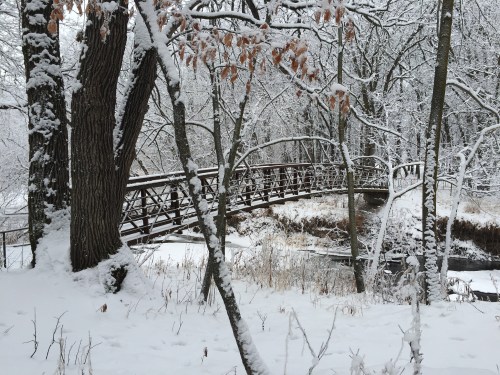 A snow-covered landscape featuring a wooden bridge surrounded by trees and a frozen stream.