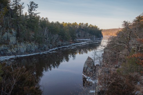 A serene river flows through a rocky landscape, surrounded by trees and reflecting the soft light of dusk.