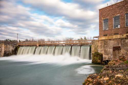 A serene waterfall cascades over a dam, with a brick building beside it under a cloudy sky.