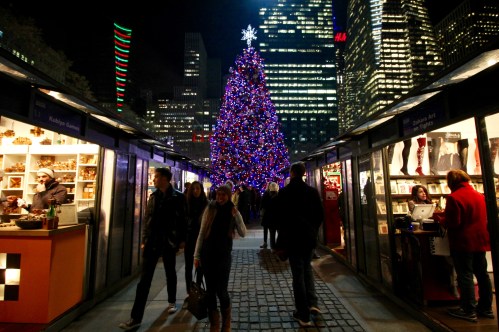 A festive scene with a large Christmas tree adorned with lights, surrounded by holiday market stalls and people walking.