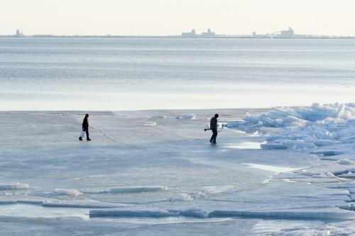 Two people walking on a frozen lake, with ice formations and a distant shoreline under a clear sky.