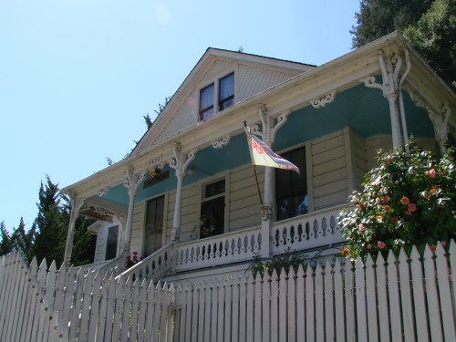 A charming two-story house with a blue roof, white trim, and a picket fence, surrounded by greenery and flowers.
