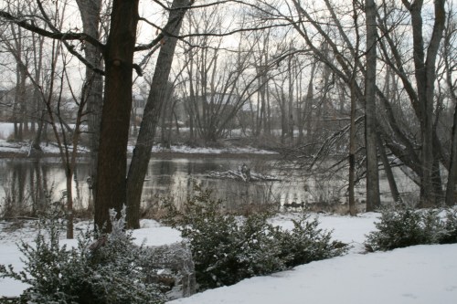 A serene winter scene with a snowy landscape, bare trees, and a calm river reflecting the overcast sky.