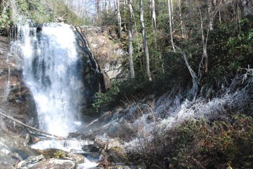 A cascading waterfall surrounded by trees and rocky terrain, with some icy patches on the ground.